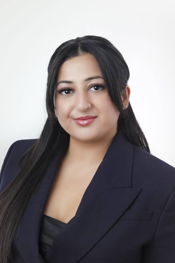 A woman with long dark hair, wearing a dark blazer and a black top, poses against a plain white background. She has a small nose ring and is smiling gently at the camera. - Jodi O Photography