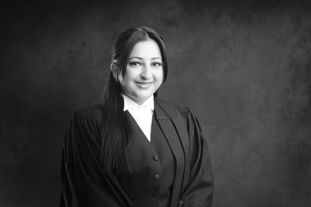 A woman in a barrister’s robe and white collar smiles at the camera against a dark, textured background in a black-and-white portrait. - Jodi O Photography