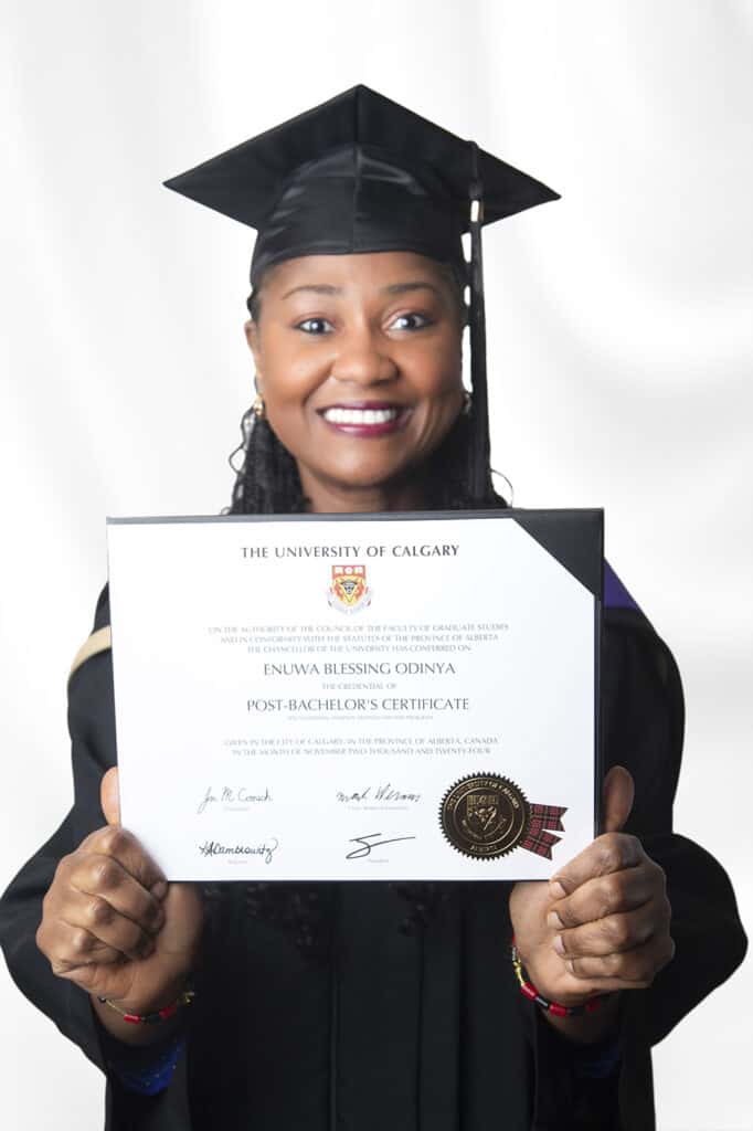 A smiling woman in graduation attire holds a University of Calgary certificate toward the camera, celebrating her achievement against a plain white background. - Jodi O Photography