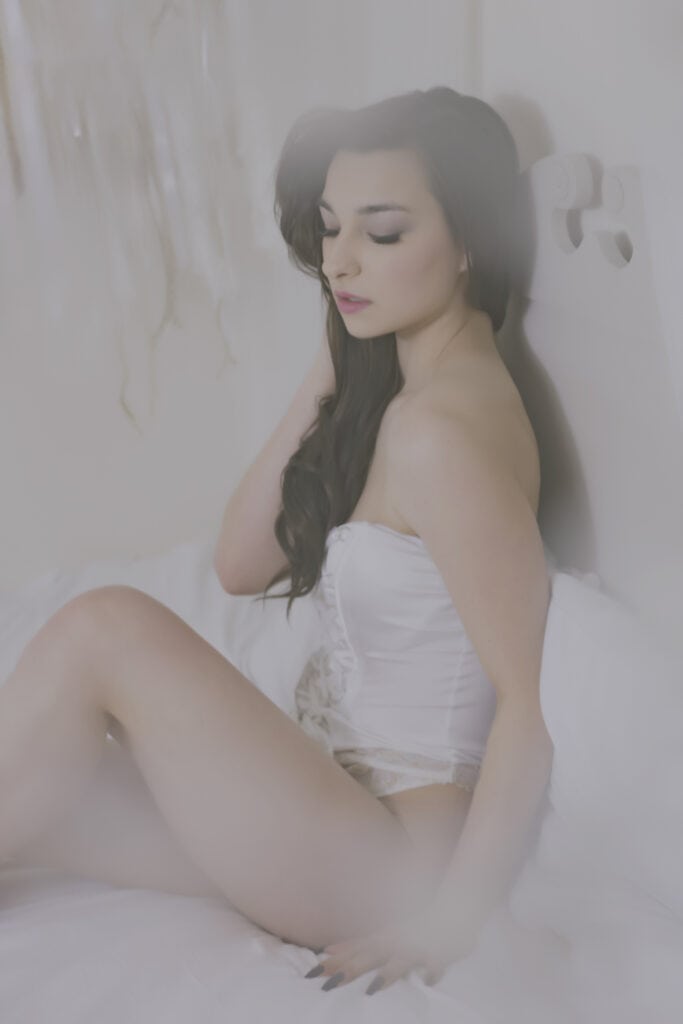 A woman with long dark hair sits on a white bed, wearing a white corset and lace underwear. She looks down with a soft, serene expression, gently touching her hair. The image has a soft, dreamy, and ethereal quality. - Jodi O Photography