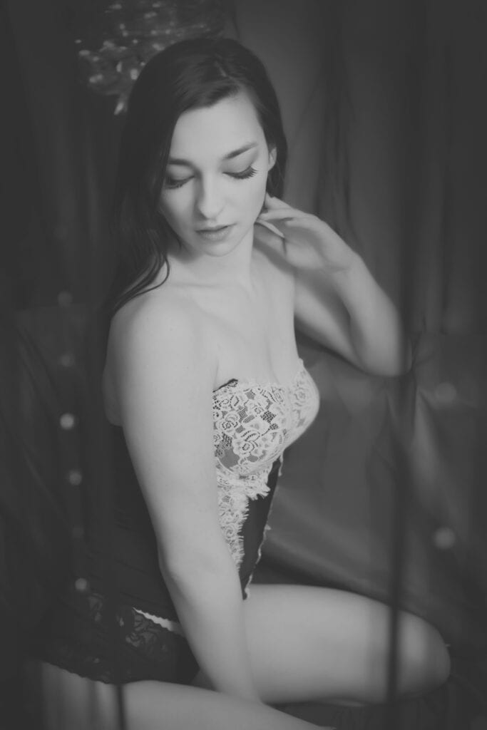 Black and white photo of a woman in lace lingerie, sitting with her legs folded, looking down and gently touching her neck; an elegant Calgary Boudoir Photographer captures her soft expression against draped fabric. - Jodi O Photography