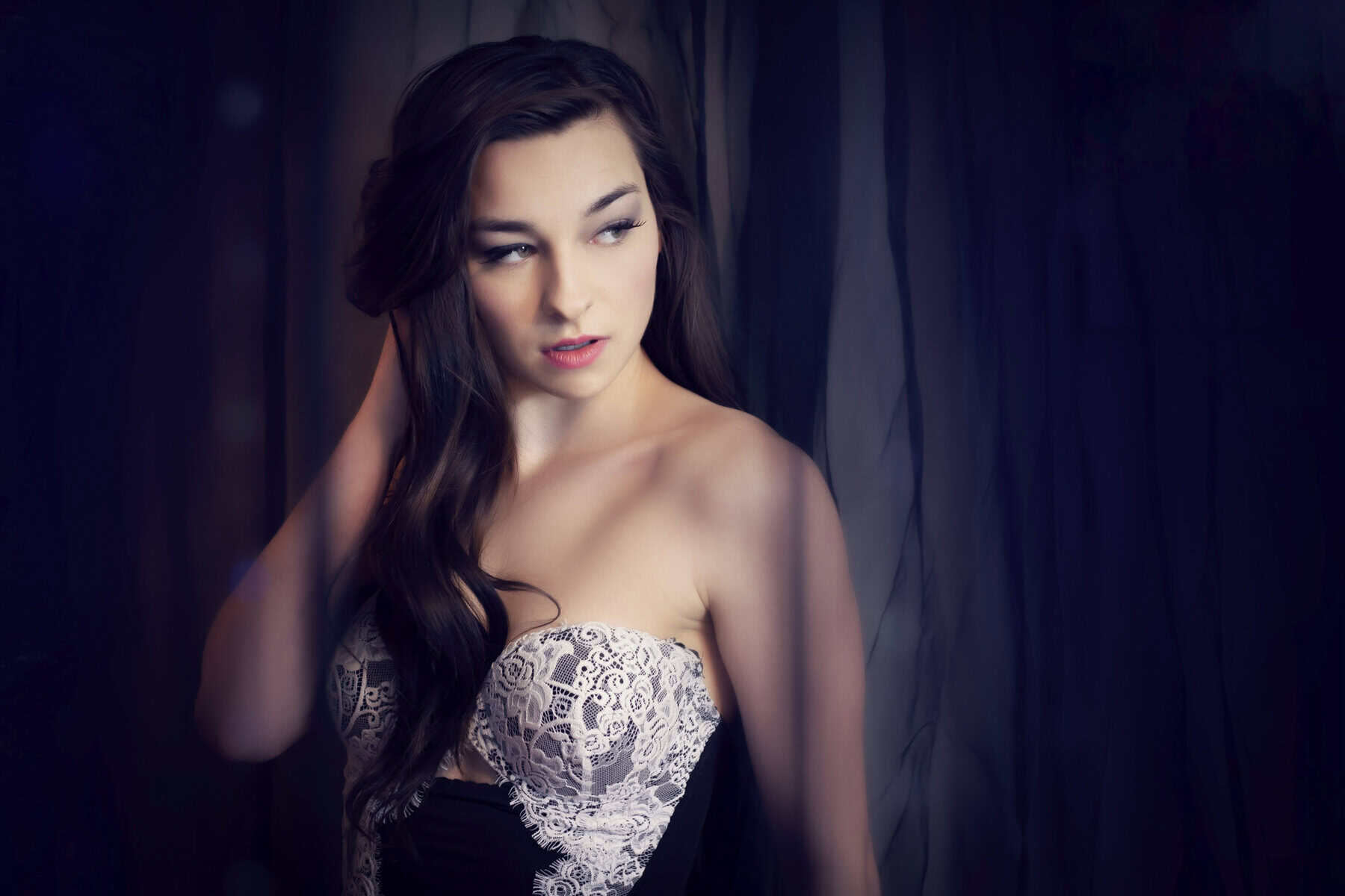 A woman with long, dark hair wearing a strapless dress with a white lace bodice stands against a dark, softly lit background. Captured by a Calgary Boudoir Photographer, she gazes to the side with her hand touching her hair. - Jodi O Photography