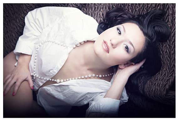 A woman with dark hair, wearing a white shirt and a long pearl necklace, lies on a textured brown surface, gazing up at the camera with a relaxed and serene expression. - Jodi O Photography