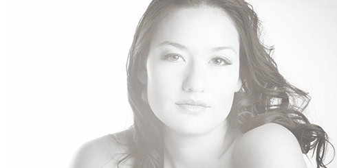A softly lit, high-key portrait of a woman with long wavy hair, looking gently at the camera with a calm expression against a white background. - Jodi O Photography