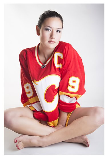 A woman sits cross-legged on the floor, wearing a red Calgary Flames hockey jersey with the number 9 and a captain's "C" on it, against a plain white background. - Jodi O Photography