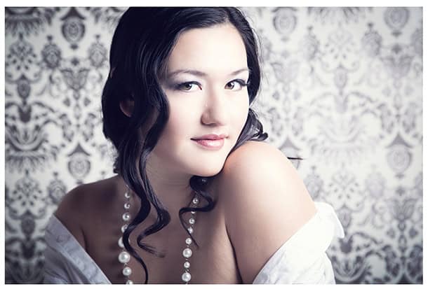 A woman with dark wavy hair and a pearl necklace poses with her shoulder exposed, wearing an off-shoulder white garment, in front of an ornate, patterned background. - Jodi O Photography