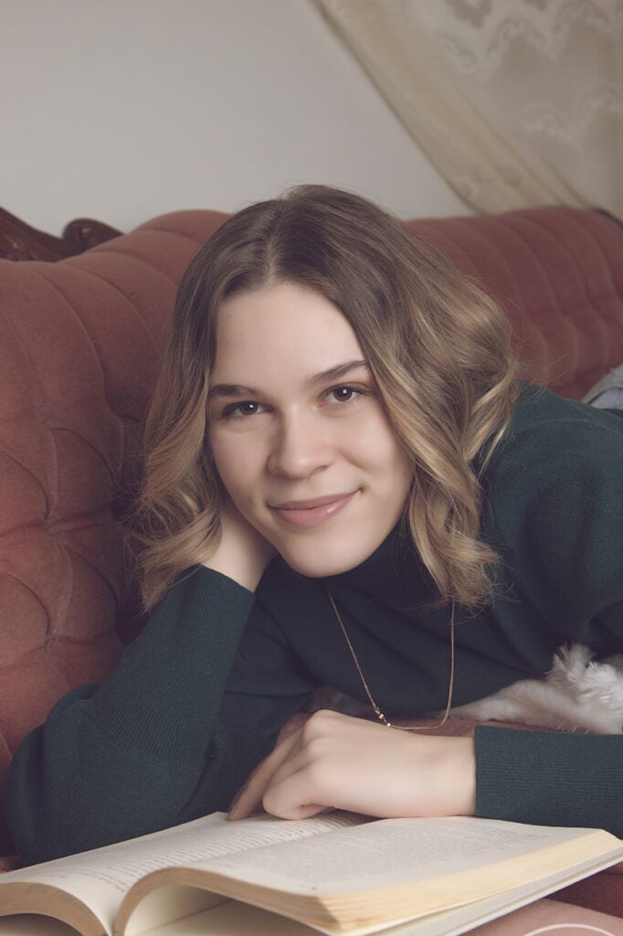 A young woman with wavy, shoulder-length hair lies on a pink couch, smiling gently at the camera. She rests her head on her hand and has an open book in front of her. - Jodi O Photography