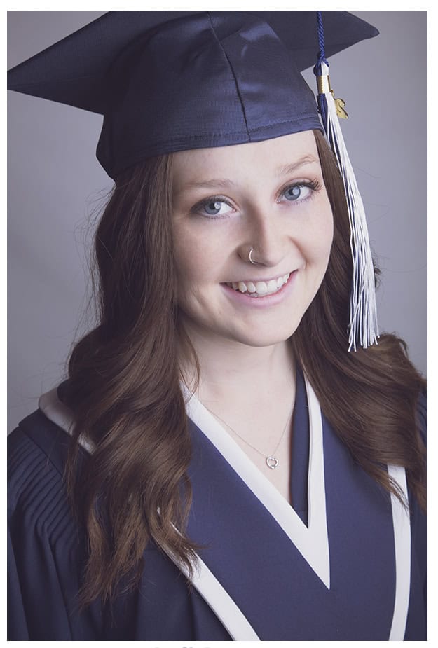 A young woman with long brown hair wearing a navy blue graduation cap and gown smiles at the camera. She has a nose ring and a heart-shaped necklace, and a white tassel hangs from her cap. - Jodi O Photography