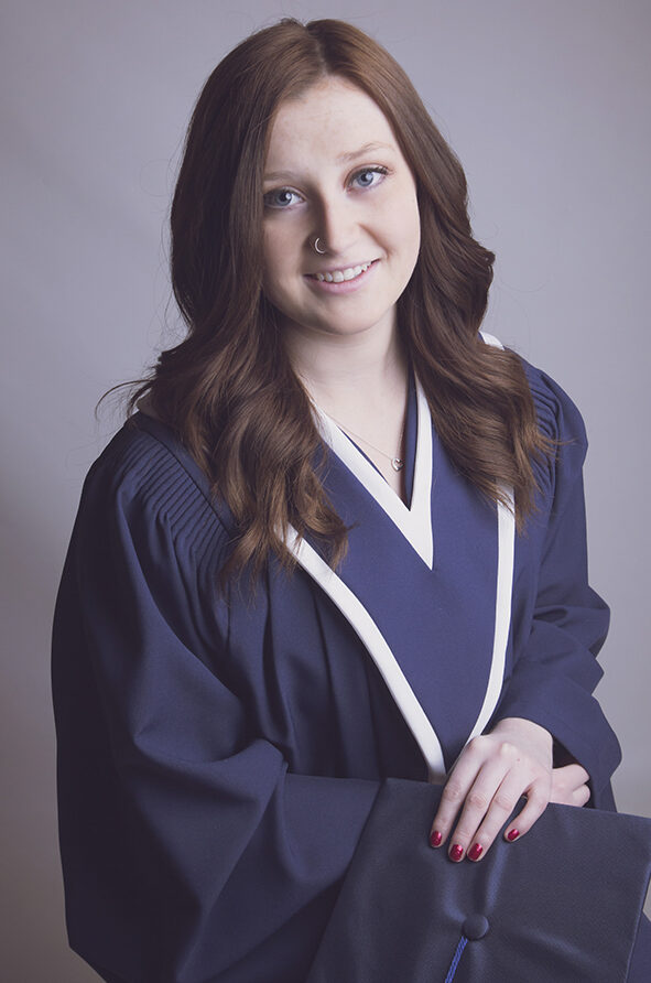 A young woman with long brown hair wearing a navy blue graduation gown holds a matching cap and smiles at the camera against a light gray background. - Jodi O Photography