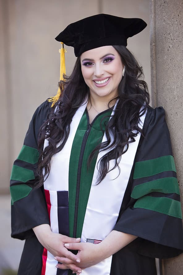 A smiling woman in a graduation gown and cap with a yellow tassel stands against a wall, wearing a white stole with green and black accents. - Jodi O Photography