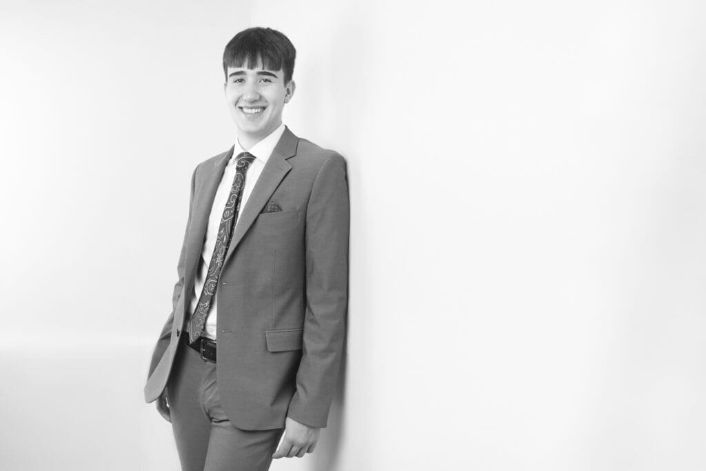 A young man in a suit and tie stands smiling, leaning against a white wall in a bright, minimalist setting. The image is in black and white. - Jodi O Photography
