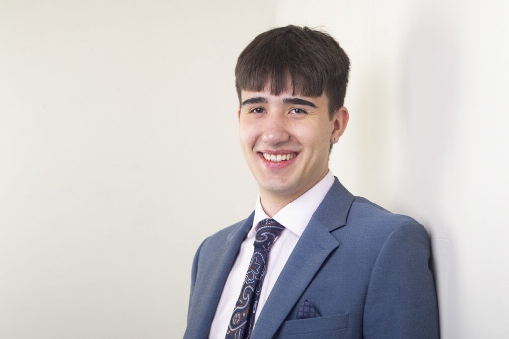 A young man in a blue suit, white shirt, and patterned tie stands smiling against a plain light-colored wall. He has short dark hair and is wearing an earring. - Jodi O Photography