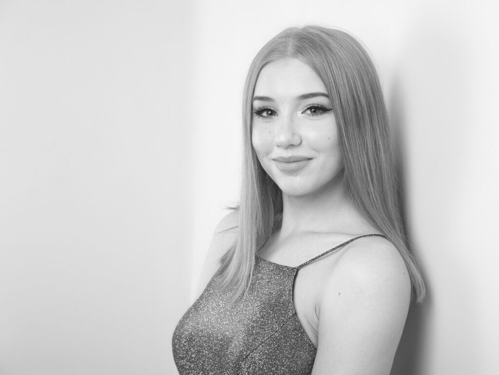 Black and white photo of a young woman with straight, light hair, wearing a sparkly sleeveless top, standing against a wall and smiling at the camera. - Jodi O Photography