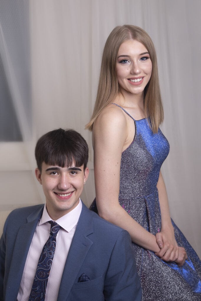 A young man in a blue suit sits beside a young woman in a sparkly blue dress. Both are smiling at the camera against a neutral indoor background with soft lighting. - Jodi O Photography