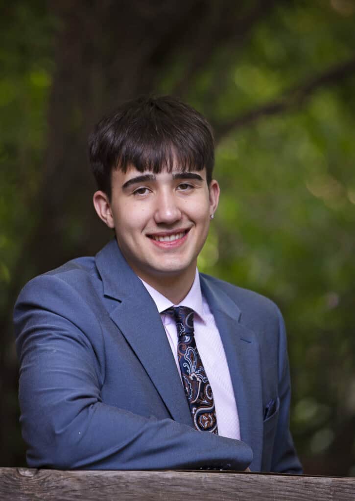 A young man in a blue suit and patterned tie smiles at the camera while leaning on a wooden railing, with a blurred green leafy background behind him. - Jodi O Photography