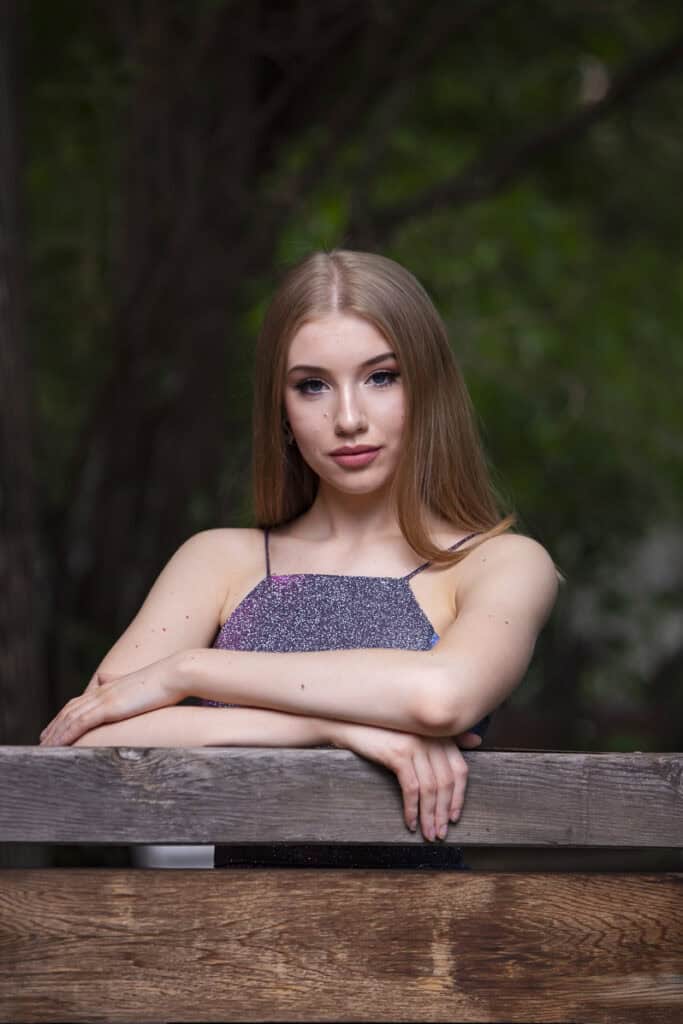 A young woman with long, straight blonde hair and a shimmery sleeveless top stands outdoors, leaning on a wooden fence with her arms crossed, surrounded by green foliage in the background. - Jodi O Photography
