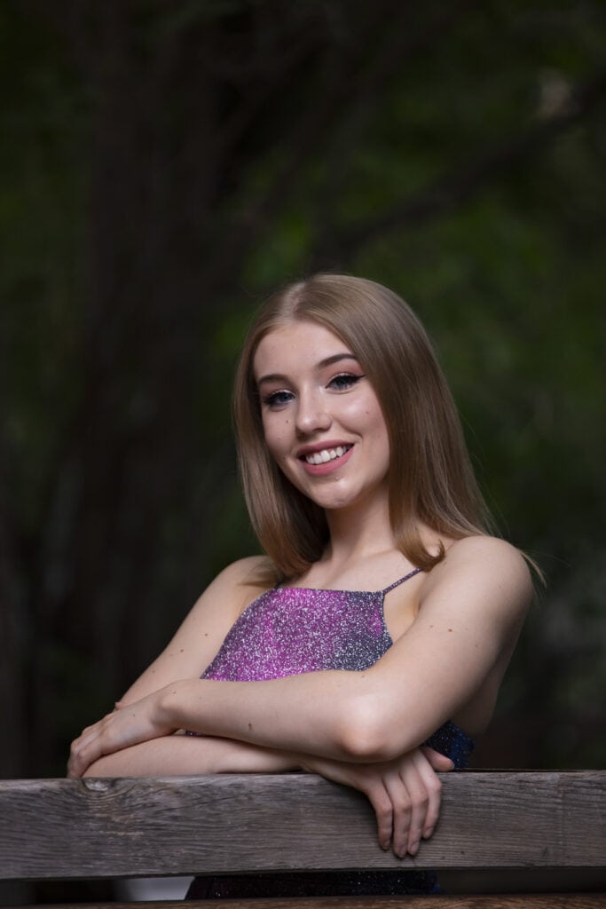 A young woman with long blonde hair smiles while leaning on a wooden railing, wearing a sleeveless, sparkly pink and purple top, with a blurred green forest background. - Jodi O Photography