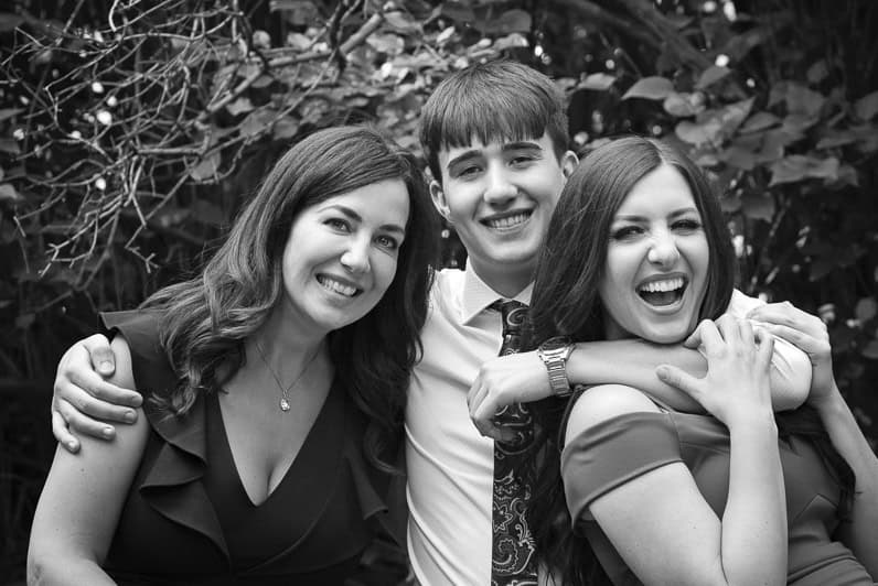 Three young adults, two women and one man, stand close together outdoors, smiling and laughing. The man has his arms around the women, and leafy trees fill the background. The photo is in black and white. - Jodi O Photography