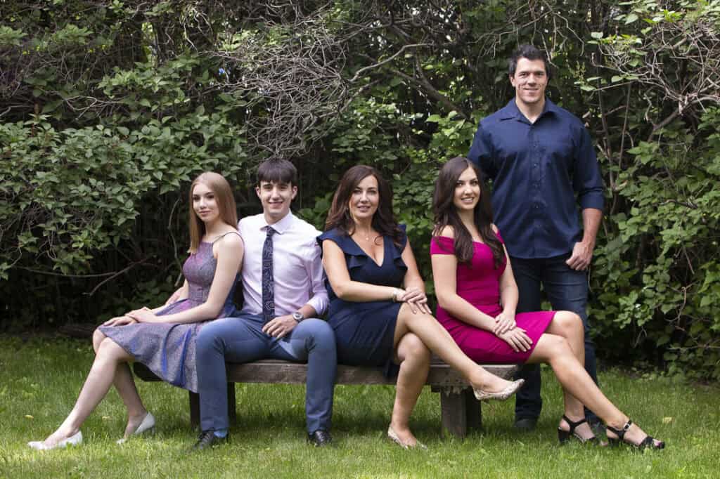 Five people pose outdoors in front of leafy bushes; four sit on a wooden bench, dressed in semi-formal attire, while one man stands behind them, smiling. The scene is bright with green grass and trees. - Jodi O Photography